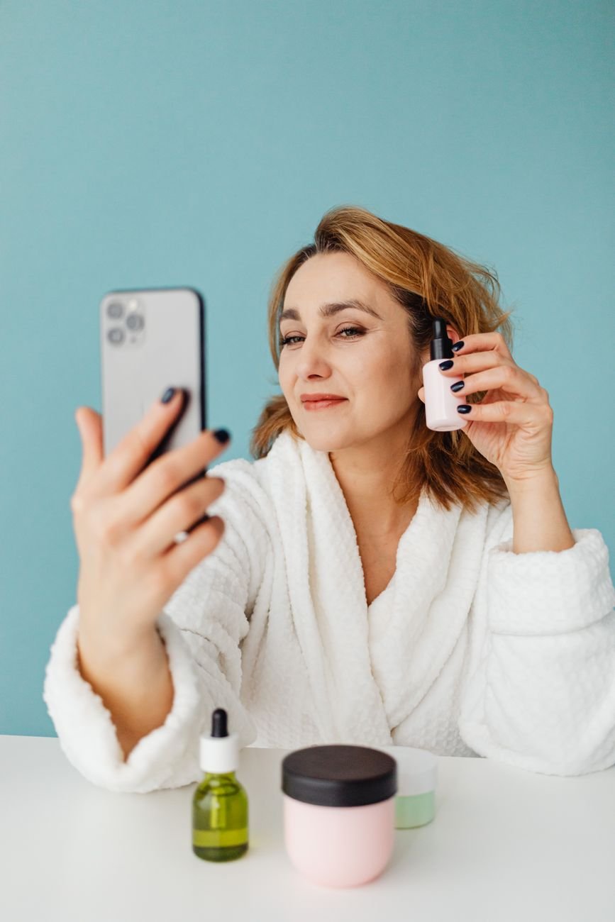 Woman in a white robe taking a selfie with skincare products on table.